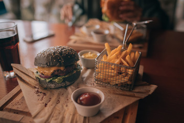 Hands holding fresh delicious burger with french fries, sauce and beer on the wooden table top view. Tasty grilled beef burger with lettuce and mayonnaise served on pieces of brown paper on a table.