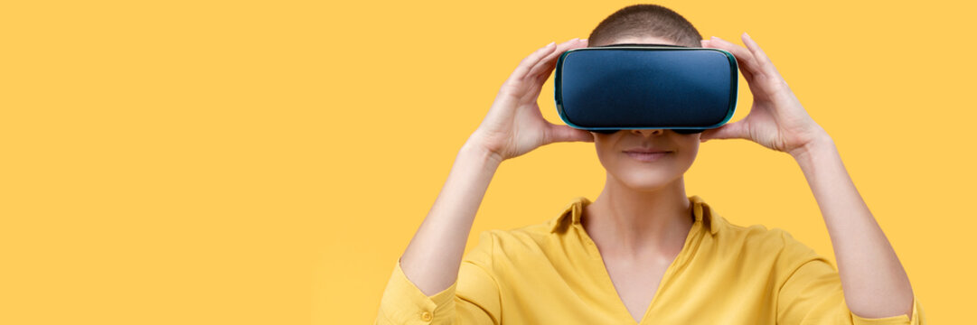 Young woman in her 30s using virtual reality goggles. Woman wearing VR headset isolated over yellow banner. VR experience concept.