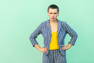 Portrait of serious handsome beautiful short hair young stylish woman in casual striped suit standing with hands on wasit and looking at camera. indoor studio shot isolated on light green background.