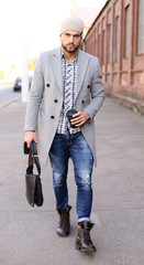 City life. Stylish young man in grey coat and hat walking on the street in the city.