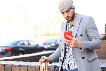 Handsome young man in grey coat and hat using smartphone, resting, standing with longboard. Urban skateboarding concept.
