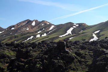 View over field with black volcanic lava ash on mountain range with spots of snow and ice contrasting with blue cloudless sky - Landmannalaugar, Iceland