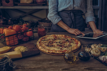 Cooking, food, technology and people concept. Young woman using a tablet computer to cook pizza in her kitchen. A woman searching the cooking pecipe and preparing food ingredient before cooking.
