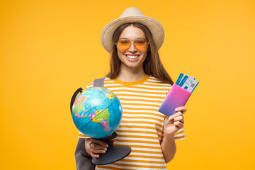 Cheerful girl holding globe in one hand and passport with flight tickets in other, isolated on yellow background