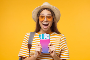Happy surprised young female tourist  holding passport with flight tickets, isolated on yellow...