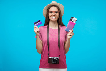 Young female tourist holding credit card and passport with tickets, ready to flight, isolated on blue background - Powered by Adobe