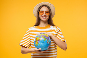 Worldwide tourism concept. Young female tourist holding globe with both hands, isolated on yellow...