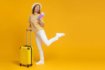 Happy female tourist jumping, holding suitcase and passport, flight tickets, isolated on yellow background with copy space