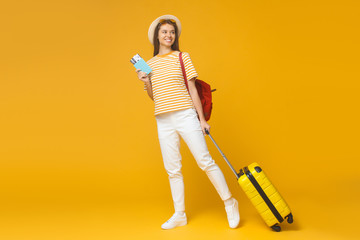 Smiling tourist girl standing with suitcase and backpack, isolated on yellow background. Travel concept