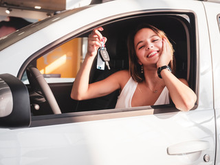 Fototapeta na wymiar Side view of a beautiful girl smiling and looking at camera. Gorgeous young woman holding keys of her new white auto. Happy female client of car dealership buying automobile