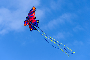 Colorful red yellow butterfly shaped kite with long tail flying in the blue sky among the clouds -...