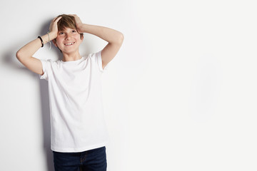 Concept of children style and fashion. Smiling young boy in white T-shirt posing in front of white empty wall background. Mockup