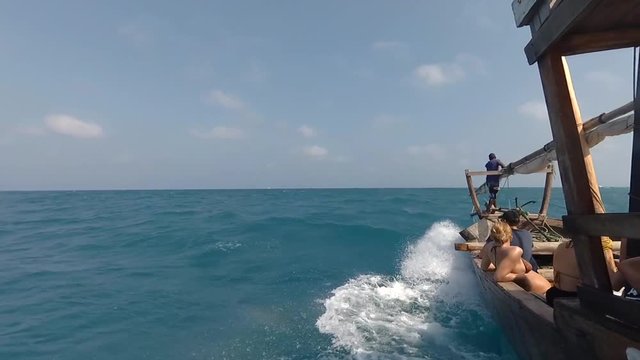 Traditional wooden Dhow boat with tourists sailing fast through ocean waves on bright sunny day. Sea horizon with tropical island and blue sky in the background. View from the left side of the boat.