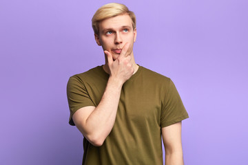 Handsome serious puzzled young man looking up with thoughtful and skeptical expression,solving a aproblem, holding finger on his chin,isolated over violet background
