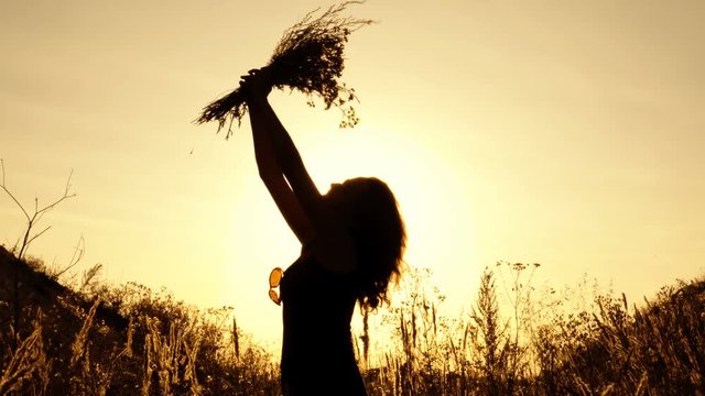The girl holds a bouquet of wildflowers at sunset in the open air, sniffs it and raises it up. Silhouette. Close up. 4K.