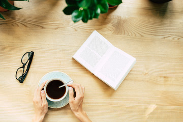 Happy beautiful young woman in glasses is reading book and drinking coffee in modern cafe