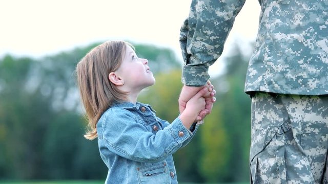 Little girl holding hand of her father with both hands. Daughter looking at her soldier father.