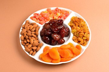 Mix of dried fruits and nuts on a white plate. Apricot, almond, raisin, dates fruit. On a brown background. Space for text or design.