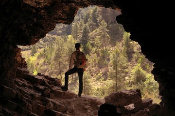 MAN ADVENTURER MOUNTAIN MAN WITH BACKPACK IN A CAVE IN FRONT OF A VALLEY FULL OF GREEN TREES