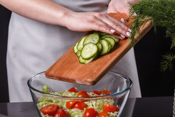 A young woman in a gray apron adds a cucumber to a salad