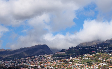 a panoramic view of funchal in Madeira with houses and buildings against the tree covered mountains with blue sky and clouds