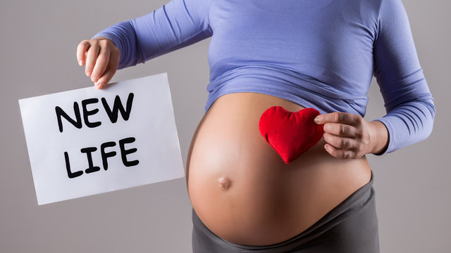 Image of close up stomach of pregnant woman holding paper with text new life and heart shape on gray background.