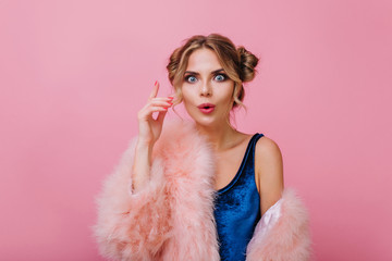Close-up portrait of surprised curly girl in velvet clothes funny posing with hand up. Amazing blonde young woman with trendy make-up remembered something important, isolated on pink background.