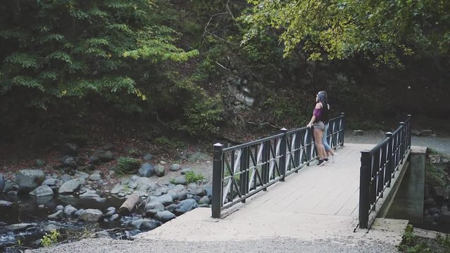 two ladies standing on bridge over fast bubbling water in Borjomi Park in Georgia, girl with blond hair shows brunette on sight with finger, travelers walking and amissing with nature of Asian country