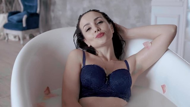dark haired lady wearing luxurious blue bra poses taking foamy bath with flower petals closeup slow motion