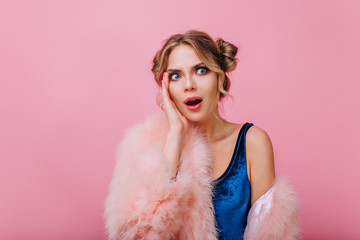 Unpleasantly surprised girl in fluffy coat and blue bodysuit looking away standing in front of pink background. Charming young woman with stylish make-up and shocked face expression touching her cheek
