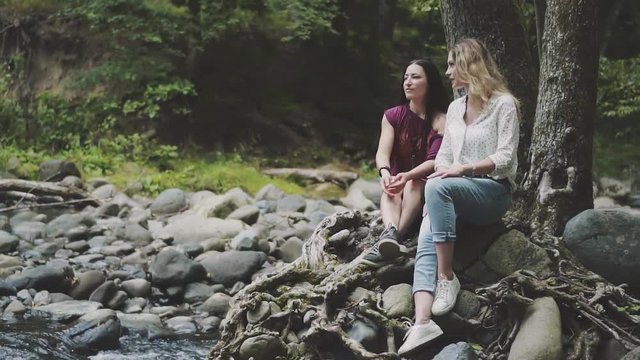 meeting of two friends on vacation, pretty young women sit on the stone shore of a mountain river, chatting and laughing with funny stories, blonde and brunette enjoy the nature of the forest