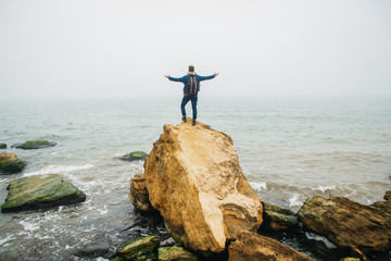 Traveler with a backpack stands on a rock against a beautiful sea with waves, a stylish hipster boy posing near a calm ocean during a wonderful journey around the world. Shoot from the back