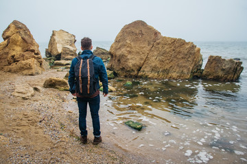 Traveler with a backpack stands on a rock against a beautiful sea with waves, a stylish hipster boy posing near a calm ocean during a wonderful journey around the world. Shoot from the back