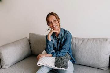 Obraz premium Smiling caucasian woman resting at home on cosy sofa at modern apartment wearing denim shirt and jeans 