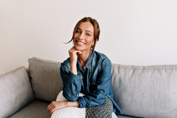 Happy laughing girl with tanned skin and white teeth sitting at sofa at cosy apartment and posing...