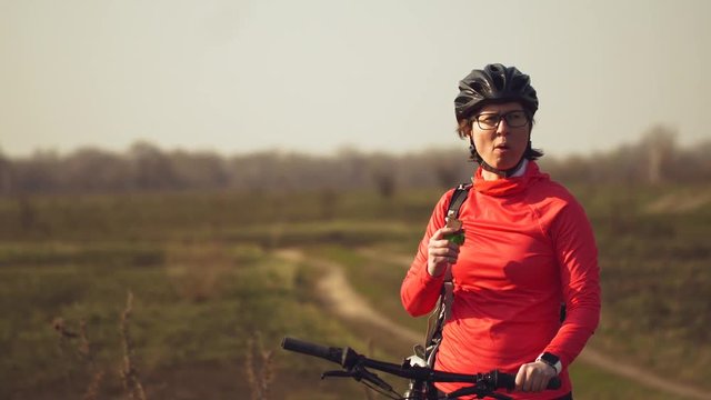 Athletic Caucasian woman eats protein bar ride on mountain bike on nature. Young sporty woman athlete in helmet resting while biting nutritional bar. Fitness woman eating energy snack outdoor