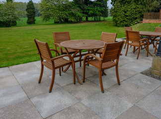 Wooden table and chairs set on a slabbed patio area with beautiful grass and tree background view.