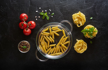 Different kinds of raw pasta with basil and tomatoes on black slate background. Top view.