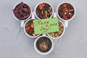 Variety of dried tea in cups on gray background. Set of aromatic dry teas and card with inscription Have a nice Day. Flat lay composition.