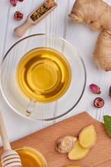 Cup of tea and seasoning, top view. Glass cup of ginger tea, ginger root, honey, dried tea and green plant on wooden background.