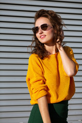 Glorious brunette woman with curly hair wearing glasses and yellow sweater posing near the shutters