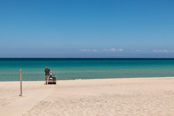 The bay of Torre dell'Orso, with its high cliffs, in Salento, Puglia, Italy. Turquoise sea and blue sky, sunny day in summer. In a bathhouse, a man cleans the sand with a small tractor (sand cleaner).