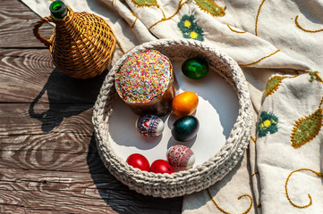 Easter still life as jug and knitted pottle with colored eggs inside stays on the agged wooden table with tablecloth under blooming cherry sprigs