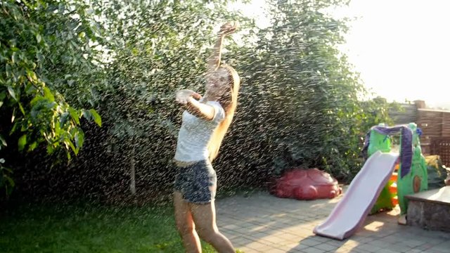 Slow motion footage of happy smiling young woman in white t-shirt dancing and jumping under warm summer rain at sunset