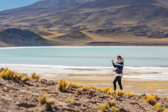 Landscape woman photographer taking mobile phone photos in an amazing wilderness environment at Atacama Desert Andes mountains lagoons. A woman cut out silhouette over the awe Tuyajto Lagoon scenery