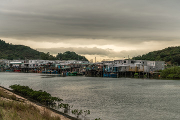 Fototapeta na wymiar Hong Kong, China - March 7, 2019: Row of Fishermen houses on stilts on Tai O River with rainy heavy cloudscape turned yellow on horizon. Sloops, tarps and simple dwellings.