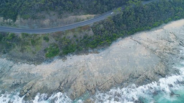 Aerial tracking shot of cars driving on narrow road near the ocean