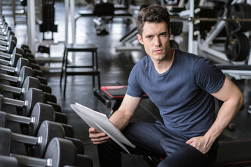 Muscular handsome trainer looking at fitness plan on clipboard for working out in the fitness gym