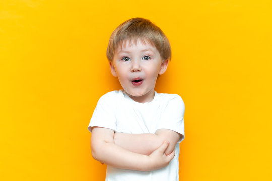 cheerful baby boy three years old in white t-shirt stands on yellow background, open his mouth surprised, blonde hair mixed race asian
