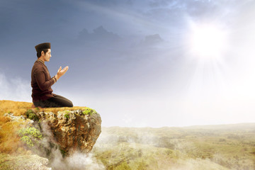 Fototapeta na wymiar Asian muslim man sitting in pray position while raised hands and praying on the edge of the cliff